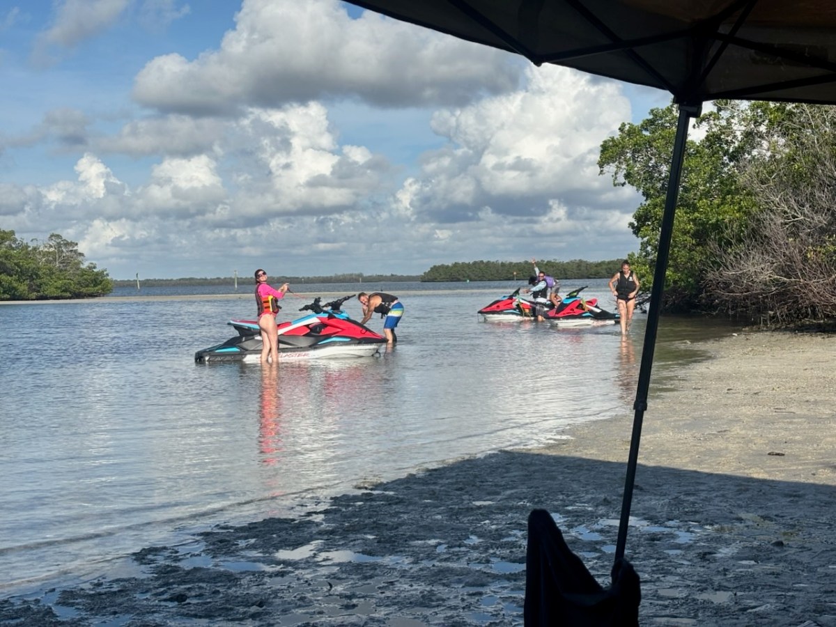 People on jet skis in shallow water near a sandy shore with cloudy sky.